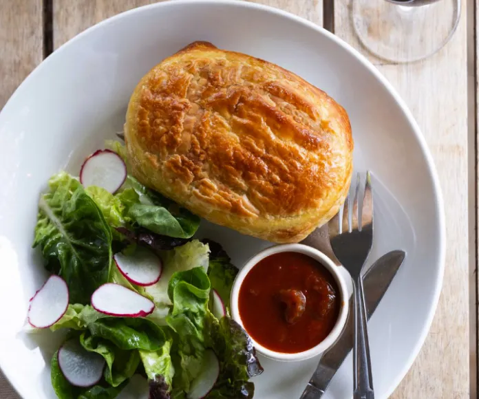 A plate from the menu features a golden-brown pastry, a side salad topped with sliced radishes, and a small cup of tomato-based dipping sauce. A fork and knife are neatly placed beside the pastry. The plate rests on a wooden surface. | Megalong Valley Tea Rooms